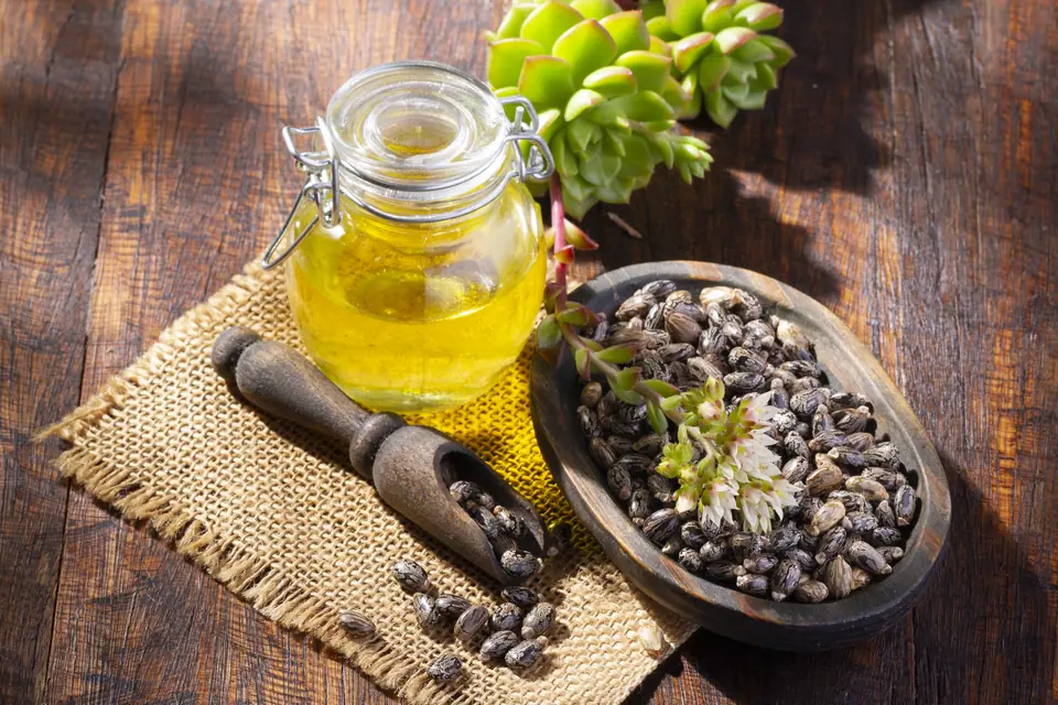 Glass jar of castor oil beside a wooden bowl of castor seeds on burlap